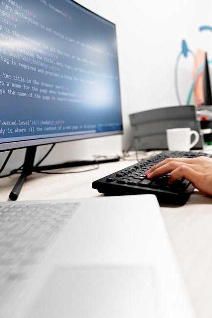 A close-up view of a person's hand operating a black keyboard in front of a desktop computer monitor displaying lines of programming code with a light blue background. The workspace includes a white d