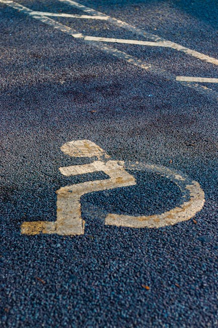 A close-up image of a designated parking space on an asphalt surface, featuring a white wheelchair symbol that indicates accessibility. The symbol shows signs of wear with faded paint and slight chipp