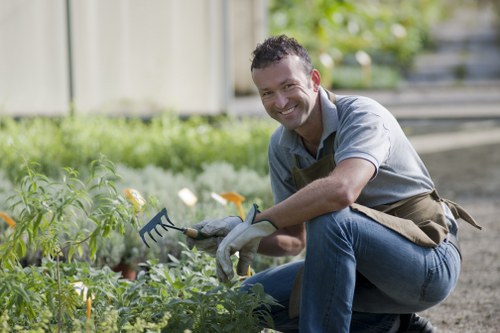 A smiling man wearing a grey shirt, brown gardening apron, and gloves is crouching in a well-maintained garden, tending to lush green plants and small flowers. The garden features a neatly edged lawn in the foreground, with a variety of plants and shrubs behind him, some with yellow and white flowers. In the background, there is a wooden fence, and the area appears to be illuminated by natural daylight, suggesting a bright, clear day. The scene depicts an outdoor landscaped space typical of a residential garden maintained by professional gardening services, with visible soil beds, leafy plants, and a paved pathway on the right side. This setting aligns with gardening and landscaping work that a company like Garden Maintenance West Ham might provide to enhance outdoor spaces in local West Ham properties.
